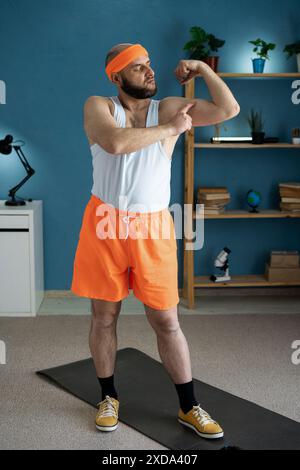 Selbstbewusster Mann, der beim Workout zu Hause den Bizeps bizepst und in orangefarbenen Shorts und Kopfbügel den Muskelfortschritt zeigt Stockfoto