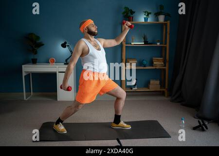 Mann, der zu Hause mit roten Kurzhanteln Ausfallschritte macht, trägt ein orangefarbenes Stirnband, ein weißes Tanktop und orangefarbene Shorts. Er konzentriert sich auf das Trainieren seiner Beine Stockfoto