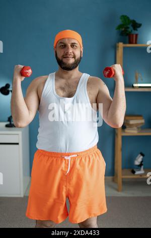 Ein Mann, der rote Kurzhanteln hob, konzentrierte sich auf das Armtraining in seinem Fitnessstudio zu Hause. Er trägt ein orangefarbenes Stirnband, ein weißes Tanktop und orangefarbene Shorts und lächelt Stockfoto