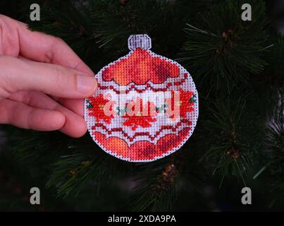 Eine Frauenhand, die ein rotes kreuzgenähtes Weihnachtsschmuck mit roten Perlen vor einem Weihnachtsbaumhintergrund hält. Stockfoto