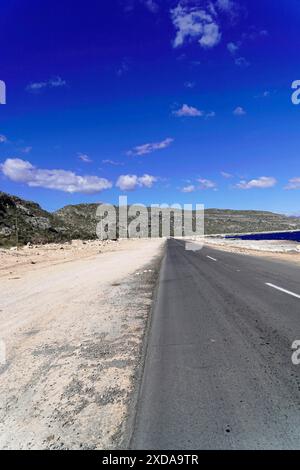 Kuba, Kuba, Zentralamerika, Eine lange, einsame Straße zieht sich durch eine weite und trockene Landschaft unter blauem Himmel Stockfoto