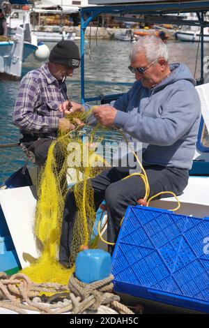 Zwei Fischer arbeiten an einem gelben Fischernetz auf einem Boot im Hafen unter freiem Himmel, umgeben von der Hafenatmosphäre und einem sonnigen Tag, Symi Stockfoto