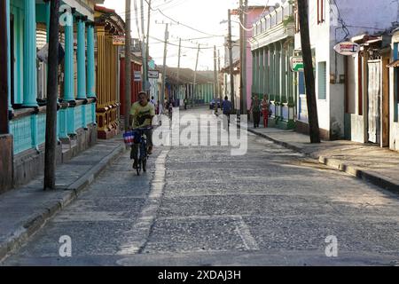 Baracoa, Kuba, Zentralamerika, Menschen und Radfahrer bewegen sich entlang einer alten Stadtstraße, die von farbenfrohen Gebäuden gesäumt ist, Kuba Stockfoto