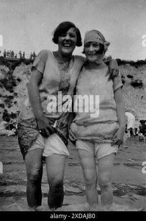 Beine und Blüten, zwei Damen im flachen Wasser, ein Strand und eine Klippe dahinter. Mode und Kultur. Aus einer Sammlung von Schnappschussfotos einer Gruppe von Frauen, 1928. Es gab nur wenige Anmerkungen auf den Fotos, aber die Frauen schienen als Gruppe zu reisen und hatten eindeutig eine Vorliebe für die englische Grafschaft Kent. Einige der Fotos deuten darauf hin, dass sie Feministinnen ihrer Zeit waren, und obwohl sie weitgehend unbekannt sind, habe ich sie aufgenommen, da sie einige der Mode der Zeit repräsentieren. Stockfoto