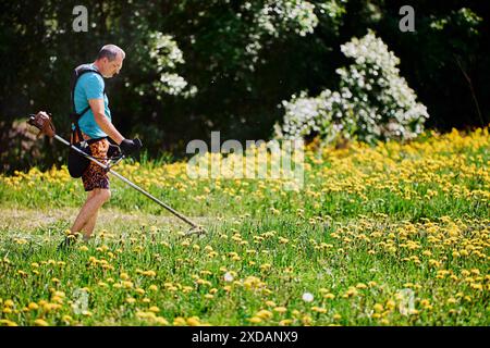 Die Rucksackaufhängung für Benzinkrauttrimmer wird von einem großen Mann getragen, der Löwenzahn auf seinem Grundstück schneidet. Stockfoto