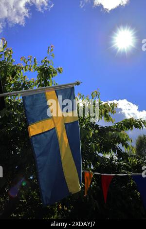 Schwedische Flagge flattert vor einem klaren blauen Himmel mit hellem Sonnenlicht und Laubbäumen im Hintergrund. Stockfoto