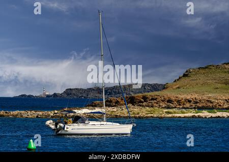 Ein Segelboot vor Anker im klaren blauen Wasser in der Nähe des Leuchtturms Faro de Favaritx auf Menorca. Die zerklüftete Küste und der dramatische Himmel bieten einen atemberaubenden Hintergrund Stockfoto