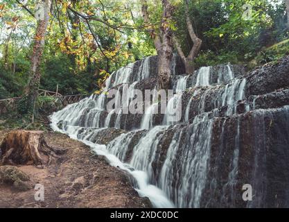 Ein ruhiger Wasserfall, der felsige Stufen hinunterstürzt, umgeben von üppigem Grün und Bäumen. Stockfoto