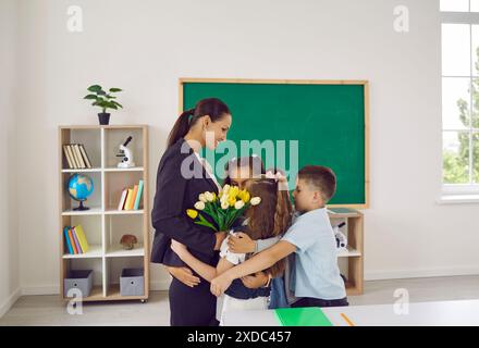 Die fröhliche Lehrerin umarmt ihre kleinen Schüler, die ihr zum Urlaub im Klassenzimmer gratulieren. Stockfoto