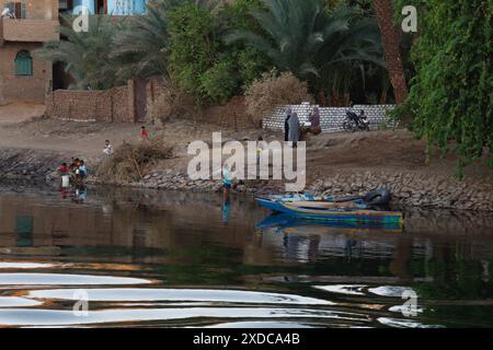 Ägyptische Frauen in farbenfroher traditioneller Kleidung unterhalten sich, während Kinder am frühen Abend am Nil in Edfu neben Ruderbooten spielen. Stockfoto