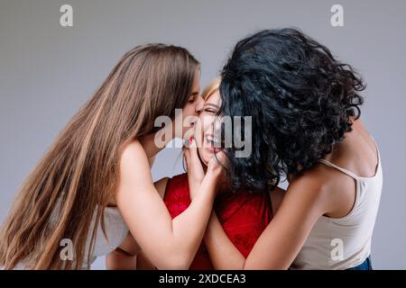 Drei Frauen küssen sich, lachen in roten Kleidern und weißen blonden Haaren, umarmen sich eng, lächeln warm und genießen die Gesellschaft des anderen Stockfoto