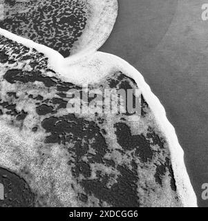 Ein Schwarzweiß-Foto, das die schwingenden schäumenden Wellen des Ozeans an einem Sandstrand zeigt. Das Bild erfasst ein dynamisches abstraktes Muster, das b erzeugt wurde Stockfoto