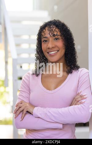 Lächelnde Frau mit lockigem Haar, die zu Hause mit gekreuzten Armen steht und selbstbewusst aussieht Stockfoto