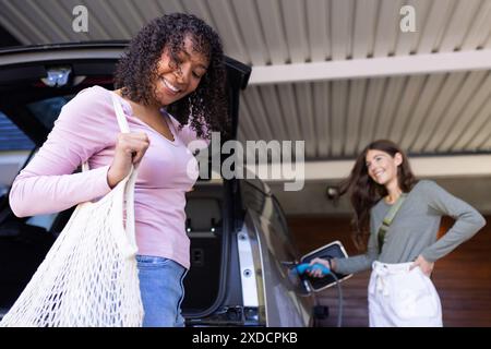 Die Frau entlädt Lebensmittel aus dem Auto, während ihr Partner ihr Elektrofahrzeug auflädt Stockfoto