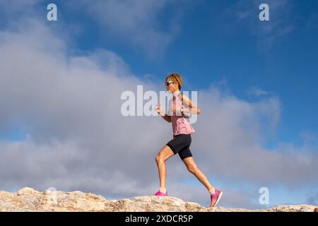 Eine athletische Frau läuft auf einem felsigen Hügel mit einem pinkfarbenen Hemd und schwarzen Shorts. Der Himmel ist blau mit ein paar Wolken. Sie ist entschlossen, den Gipfel zu erreichen Stockfoto