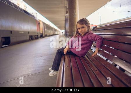 Ein kleines Mädchen in roter Jacke und Jeans sitzt auf einer Bank am Bahnhof. Ein schönes Mädchen auf dem Bahnsteig in der Nähe eines stehenden Zuges. Stockfoto