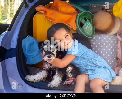 Porträt eines Kindes mit einem Schnauzer-Hund im Kofferraum eines Autos, der sich auf den Urlaub vorbereitet Stockfoto