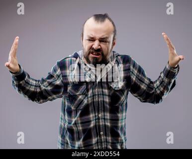 Ein bärtiger Mann drückt Frustration und Wut aus, während er seine Hände in die Luft hebt. Er ballt seine Zähne zusammen und furcht seine Stirn in einer Anzeige von n Stockfoto