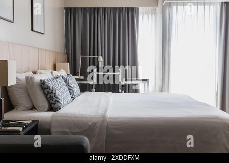 Modernes Schlafzimmer mit einem ordentlichen Bett, dekorativen Kissen, einem eleganten Schreibtisch und großen Fenstern mit Vorhängen. Ideal für Komfort, Stil und Kontinuität Stockfoto