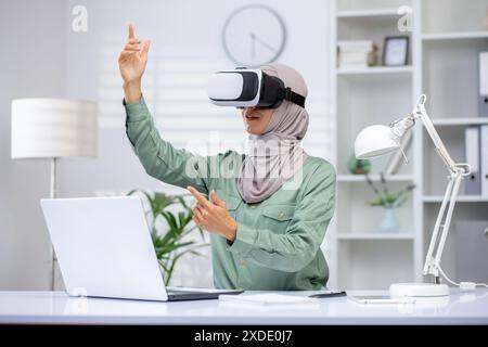 Eine junge muslimische Frau in einem Hijab sitzt im Büro an einem Schreibtisch, trägt eine virtuelle Maske und spricht fernab über Online-Kommunikation. Stockfoto