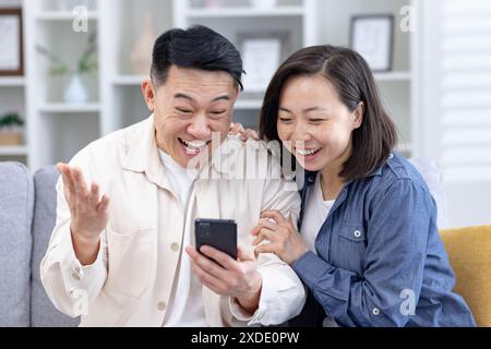 Glückliches junges asiatisches Paar, Mann und Frau sitzen zu Hause auf dem Sofa und sind zufrieden mit den Nachrichten und Nachrichten, die sie am Telefon lesen. Stockfoto