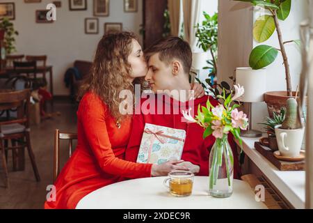Eine Frau in rotem Kleid sitzt mit einem Mann an einem Café-Tisch und küsst ihn auf die Stirn. Sie hält ein Geschenk in der Hand, eingewickelt in dekoratives Papier, und Stockfoto