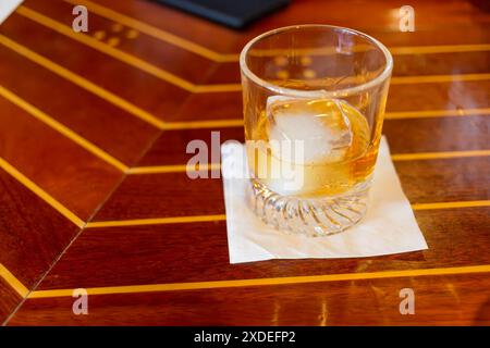 Ein Kristallglas mit köstlichem goldbraunem Bourbon und ein Eiswürfel auf einer Holzbar. Stockfoto