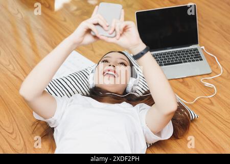 Blick von oben auf die junge schöne asiatische Frau mit Kopfhörern, die Musik vom Laptop hören und das Smartphone verwenden, während sie zu Hause auf dem Boden liegt. Stockfoto