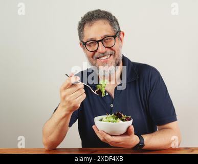 Ein Mann mittleren Alters mit Brille und Bart isst einen Salat, lächelt und blickt in die Kamera, Inneneinrichtung, gesundes Essen, Lebensstil, Freizeitkleidung Stockfoto