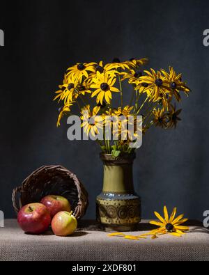 Ein Strauß gelber Gänseblümchen in einer Keramikvase und drei rote Äpfel in einem Korb auf dem Tisch. Grauer Hintergrund. Stillleben Stockfoto