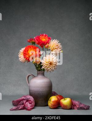 Ein heller Blumenstrauß mit Gartenblumen in einer Vase auf grauem Hintergrund, drei rote Äpfel liegen in der Nähe. Buntes Herbststillleben mit verschiedenartigen Dahlien. Stockfoto