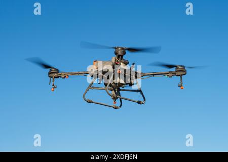 Eine Nahaufnahme einer landwirtschaftlichen Drohne mit Herbizid-Sprühgerät, die in einem klaren blauen Himmel fliegt. An der Unterseite der Drohne ist ein großer Tank angebracht, und an den Armen sind Düsen sichtbar. Stockfoto