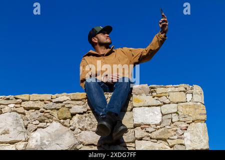 Männliches Modell, das Selfie macht, sitzt auf Steinmauer blauem Himmel Hintergrund Stockfoto