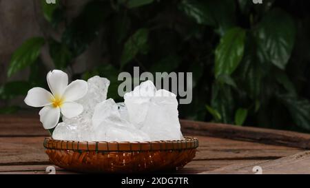 Ein Korb, gefüllt mit weißen Alaun-Kristallen, sitzt auf einem leuchtend grünen Strauchhintergrund. Die Kontrastfarben heben die Reinheit des Alaums gegenüber dem hervor Stockfoto