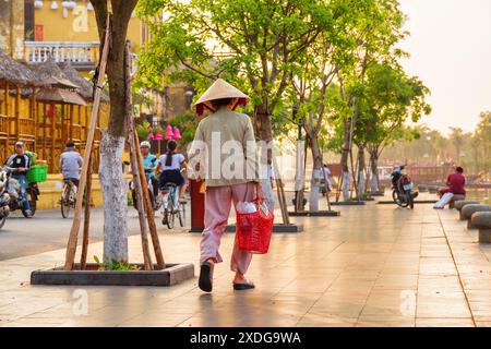 Hoi an (Hoian), Vietnam - 12. April 2018: Vietnamesische Frau mit traditionellem Bambushut, die entlang des Ufers des Flusses Thu Bon in Hoi an spaziert. Stockfoto