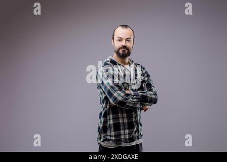 Selbstbewusster bärtiger Mann posiert mit gefalteten Armen in einem stilvollen karierten Hemd, sieht zufrieden aus und ist glücklich in Studioatmosphäre mit neutralem grauen Hintergrund Stockfoto