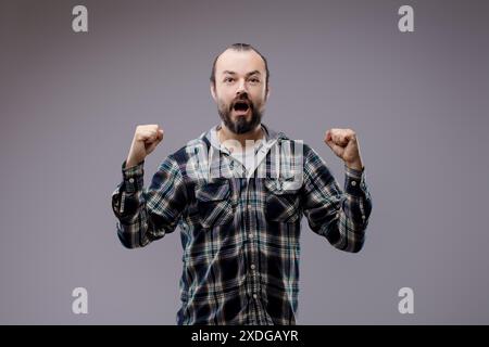 Bärtiger Mann feiert mit erhobenen Fäusten, zeigt pure Freude und Begeisterung, strahlt Erfolg und Leistung aus in einem Studio-Schuss vor grauem Hintergrund Stockfoto