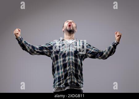 Bärtiger Mann feiert den Sieg mit erhobenen Fäusten und zeigt pure Freude und Aufregung vor grauem Atelierhintergrund Stockfoto