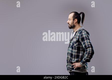 Ein bärtiger Mann in einem karierten Hemd, der vor grauem Hintergrund steht, in tiefem Denken verloren ist und Männlichkeit und Einsamkeit mit einem trendigen Hipster-Stil ausstrahlt Stockfoto