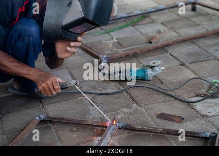 Ein Mann, der einen Schweißhelm trägt, schweißt Metallteile auf einer gepflasterten Oberfläche zusammen. Neben ihm liegt ein Elektrowerkzeug. Stockfoto