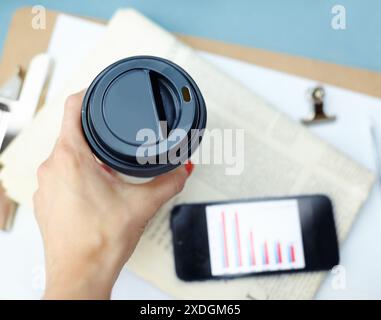 Kaffee trinken, Draufsicht und Handnaht mit Telefondaten, Zeitungen oder Informationen. Tasse, Tee oder Frau mit Getränk oben für Finanzanalytiker Stockfoto