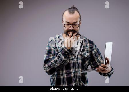 Ein fokussierter Webentwickler mit Laptop erwägt Codierungsprobleme im Studio. Grauer Hintergrund verbessert die Atmosphäre. Expertise und Entschlossenheit sind nachweisbar Stockfoto