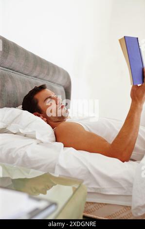 Mann, buchen und lesen Sie im Bett, um sich zu entspannen, Fantasie und Wissen am Morgen oder Wochenende. Männlicher Journalist, komfortabel und gemütlich mit Literatur und Fiktion Stockfoto
