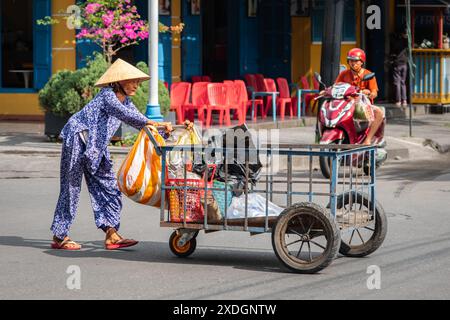 Vietnamesische alte Frau schiebt einen Wagen mit Müllsäcken. Hoi an, Vietnam. Eine Frau in traditioneller vietnamesischer Kleidung, die einen Metallwagen schiebt. Reisen Stockfoto