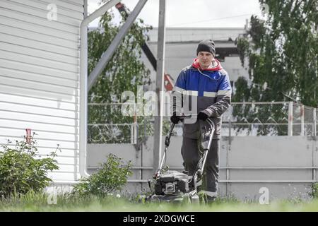 Ein Mann in einer blau-grauen Jacke mäht den Rasen. Er trägt einen Hut und Handschuhe Stockfoto