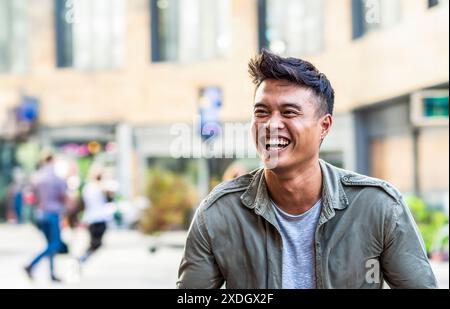 Junger asiatischer Mann lacht fröhlich in einer städtischen Umgebung mit verschwommenem Stadtbild. Das Bild fängt einen Moment echten Glücks und Spontanei ein Stockfoto