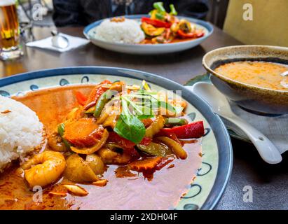 Rote Currysauce mit Garnelen und Gemüse auf zwei Tellern, Suppe in einer Schüssel und Bier auf dem Tisch. Asiatische Küche. Stockfoto