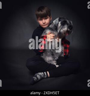 Ein Junge sitzt auf dem Boden mit einem Hundezwergschnauzer in den Armen. Der Junge trägt einen schwarzen Hoodie. Der Hund ist eine kleine, schwarze und graue Rasse Stockfoto