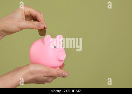 Frau, die Münze in rosa Schweinebank auf olivfarbenem Hintergrund steckt, Nahaufnahme. Leerzeichen für Text Stockfoto