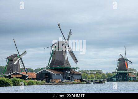 Niederlande. Ein bewölkter Sommertag in Zaanse Schans. Drei alte, aber funktionierende Windmühlen am Ufer eines Kanals Stockfoto